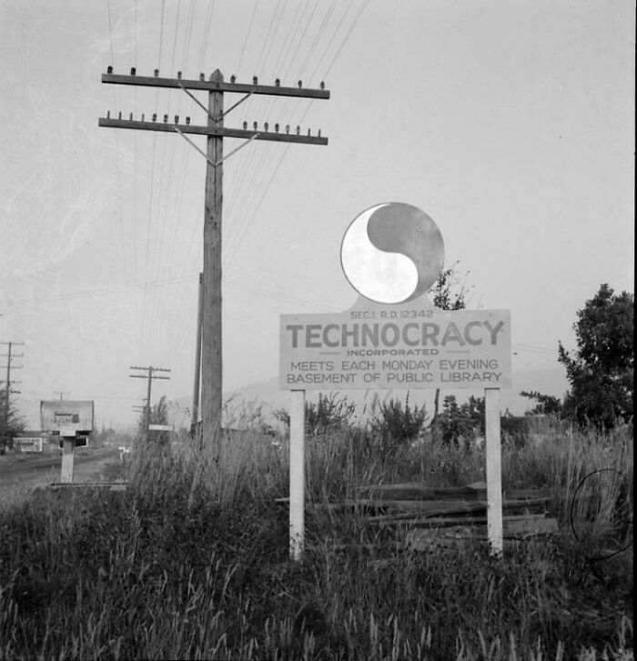 Black and white US history photo showing a technocracy meeting sign by a roadside near telephone poles and overgrown grass.