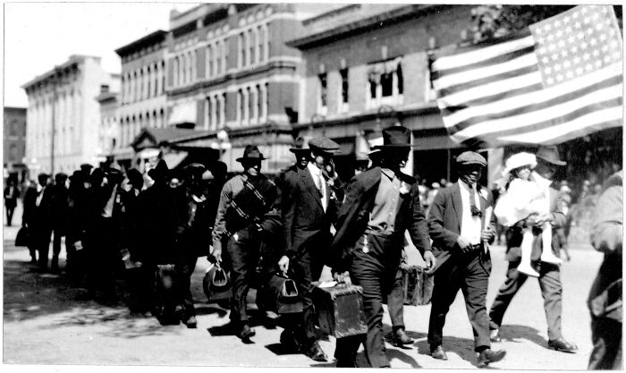 Black and white US history photo showing a line of men carrying bags and suitcases walking past buildings with an American flag.