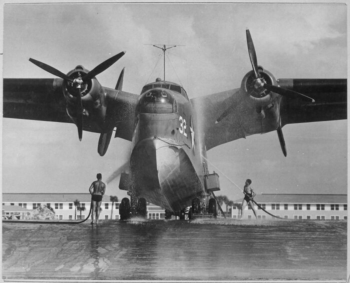 Vintage US history photo of two men washing a large military seaplane on a sunny day by a building.
