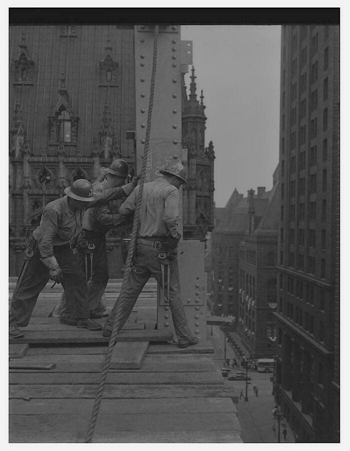 Three construction workers wearing helmets, working on steel beams high above a city street in US history photos.
