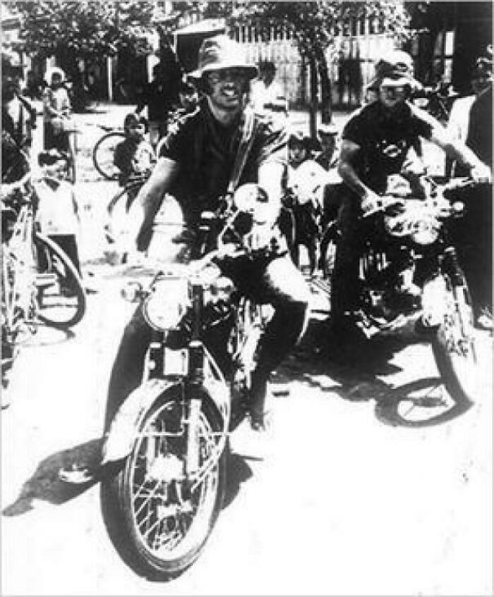 Black and white US history photo of two men riding vintage motorcycles with children watching from the street.