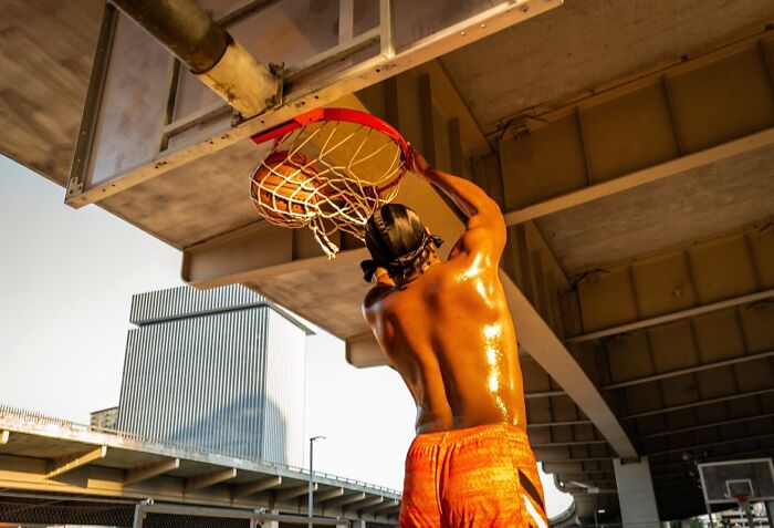 Street photo of a shirtless man dunking a basketball under an urban bridge, capturing life exactly as it happens.