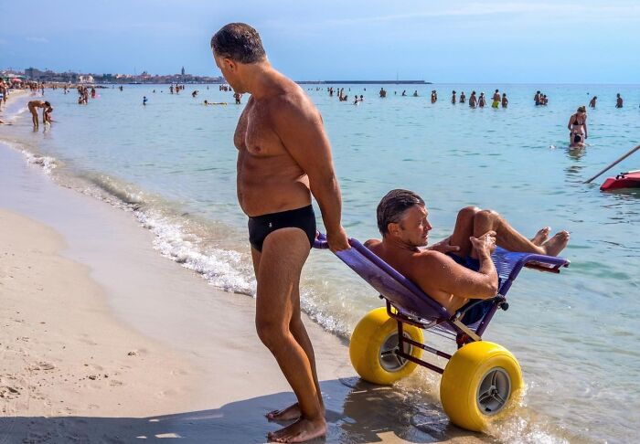 Man pushing a friend in a beach wheelchair with large yellow wheels along the shoreline in a candid street photo.