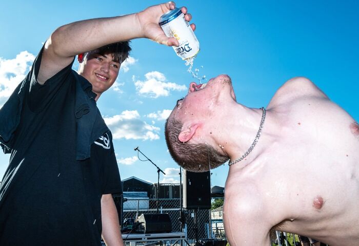 Two young men captured in a candid street photo by Perry Hall, one pouring a drink into the other's mouth outdoors.