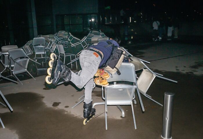 Person wearing rollerblades falling over chairs outdoors at night, a candid moment in street photos by Perry Hall.