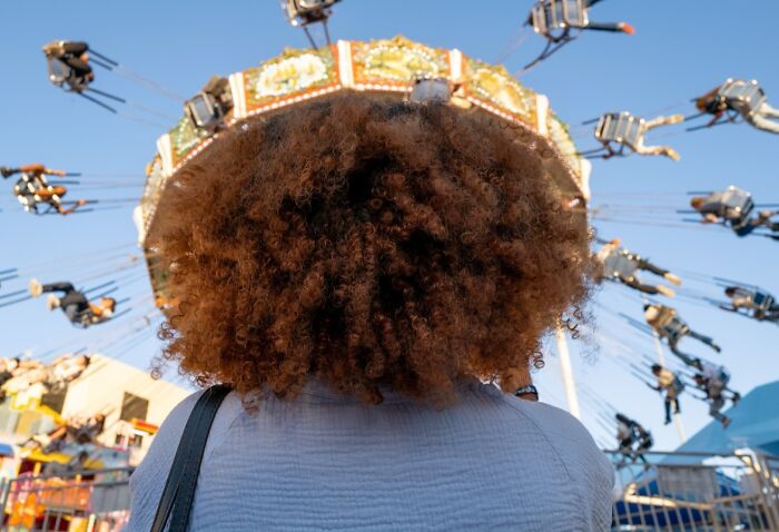 Person with curly hair viewed from behind at a busy amusement park ride, capturing life exactly as it happens in street photos.