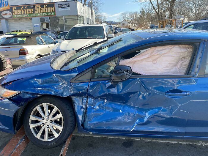 Blue car with severe side damage and deployed airbags parked outside a service station, highlighting dumb driver mistakes.