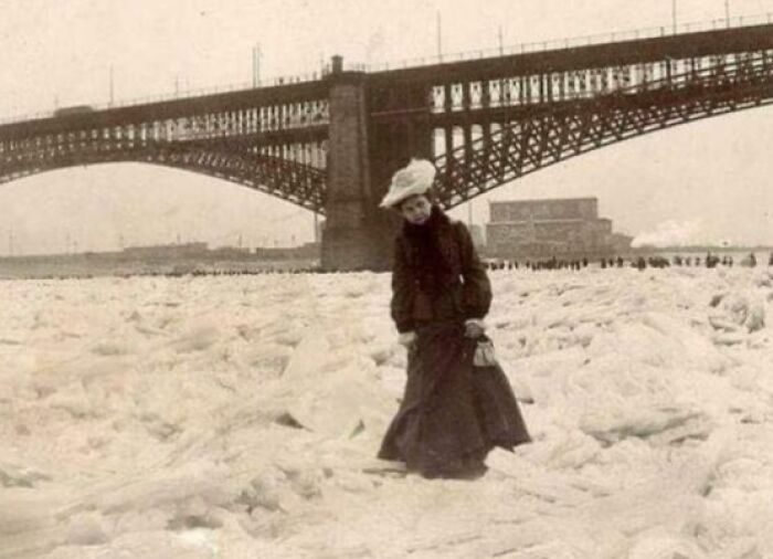 Woman in early 1900s dress standing on frozen river beneath a large city bridge in a historic US history photo.