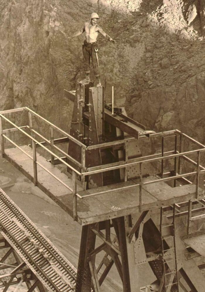 Shirtless construction worker standing atop a metal structure at a large industrial site vintage US history photo.