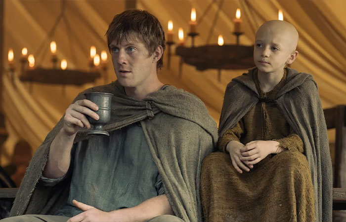 Two characters from A Knight Of The Seven Kingdoms wearing cloaks inside a candlelit medieval tent setting.