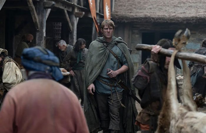 Medieval village scene from A Knight Of The Seven Kingdoms with a cloaked character walking through a busy marketplace.