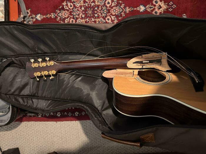 Broken acoustic guitar with shattered neck and missing strings inside a black soft guitar case, illustrating unfortunate events.