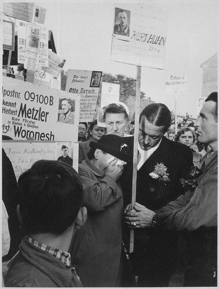 Black and white historical photograph showing a solemn crowd holding signs with portraits and names, depicting a past world.
