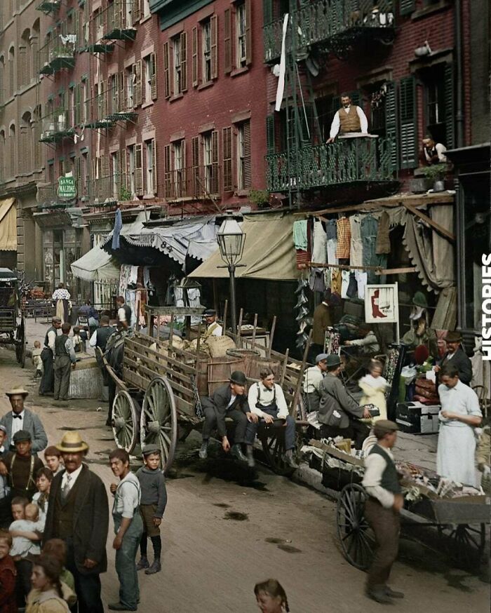 Colorized historical photograph showing a busy street scene with people, carts, and old buildings in a past world.