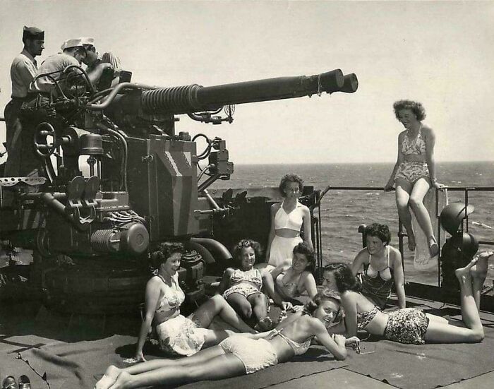 Group of women in vintage swimsuits relaxing near large naval gun on ship deck in an interesting historical photograph.