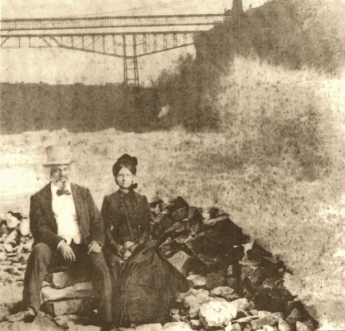 Vintage historical photograph showing a man and woman sitting by rocky shore with a bridge in the background.