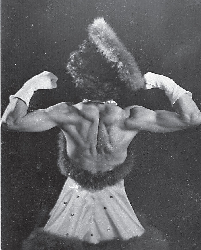 Vintage historical photograph of a muscular woman flexing her back muscles in a fur-trimmed dress and gloves.