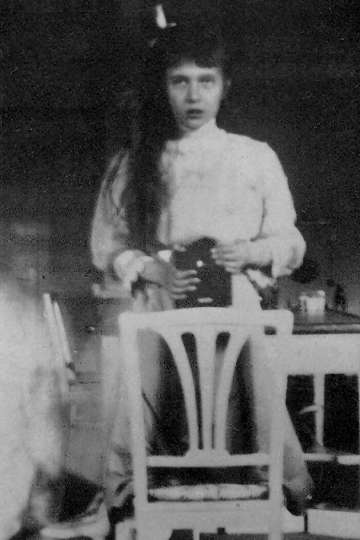 Black and white historical photograph of a woman in vintage clothing behind a wooden chair, early 1900s era.