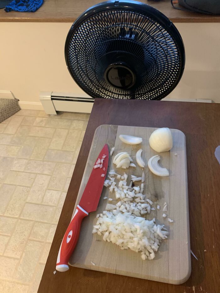 Chopped onion on a wooden cutting board with a red knife nearby and a fan blowing on the cutting area.