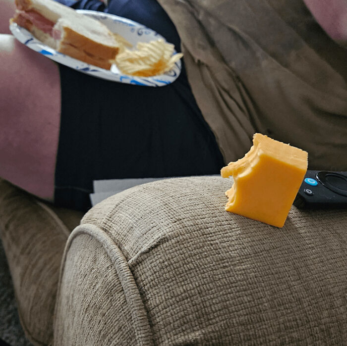 Chunk of cheddar cheese with bite taken out rests on couch arm next to remote, man lounging with sandwich and chips behind in men being men example.