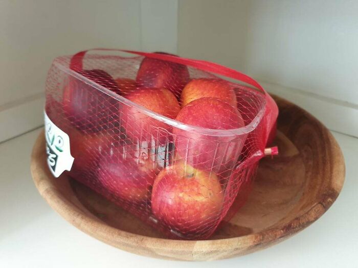 A plastic container of apples wrapped in a red mesh bag placed inside a wooden bowl, showing men being men humor.