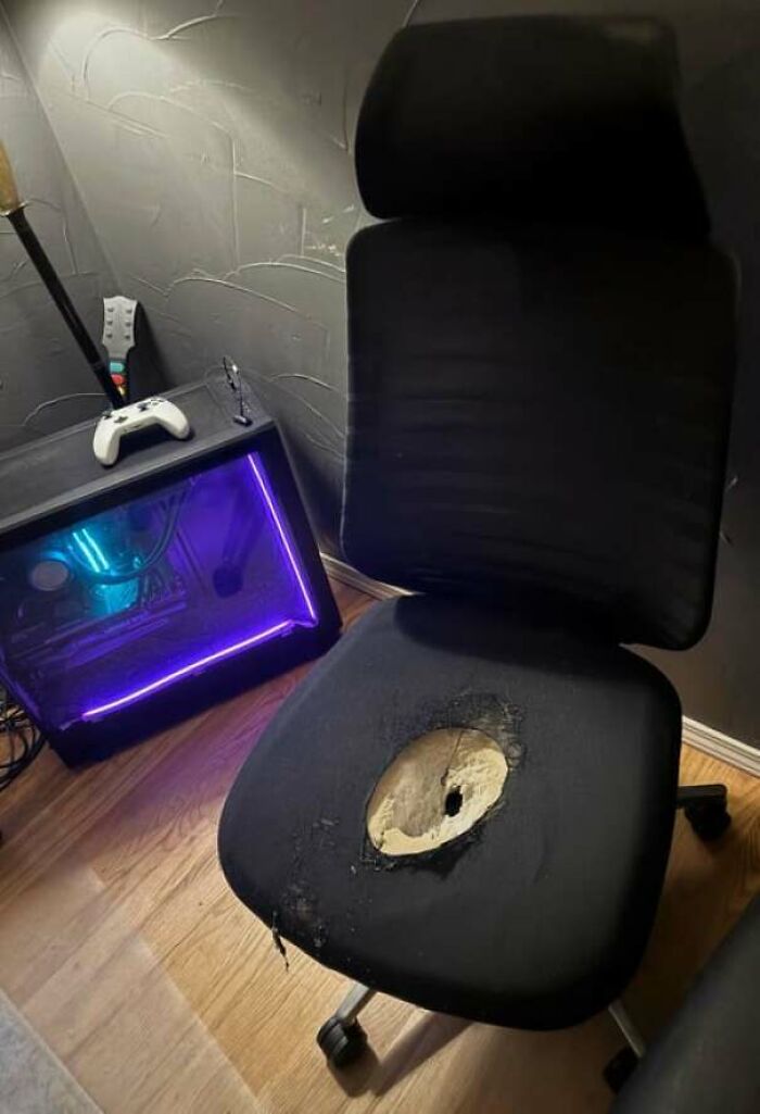 Worn-out office chair with a large hole in the seat next to a gaming PC and controllers, showing men being men humor.