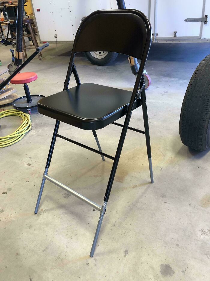 Black folding chair with extended front legs supported by metal pipe in a garage, example of if it's stupid but it works.