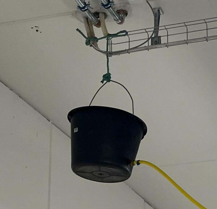 Black bucket hanging from ceiling pipes with a yellow hose attached, showing a creative if it's stupid but it works example.