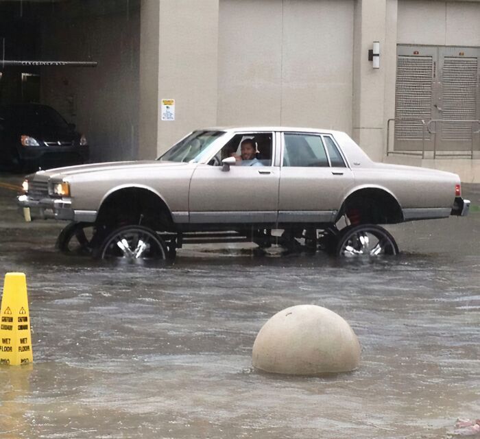 Older car modified with lifted rims driving through a flooded street, an example of if it's stupid but it works