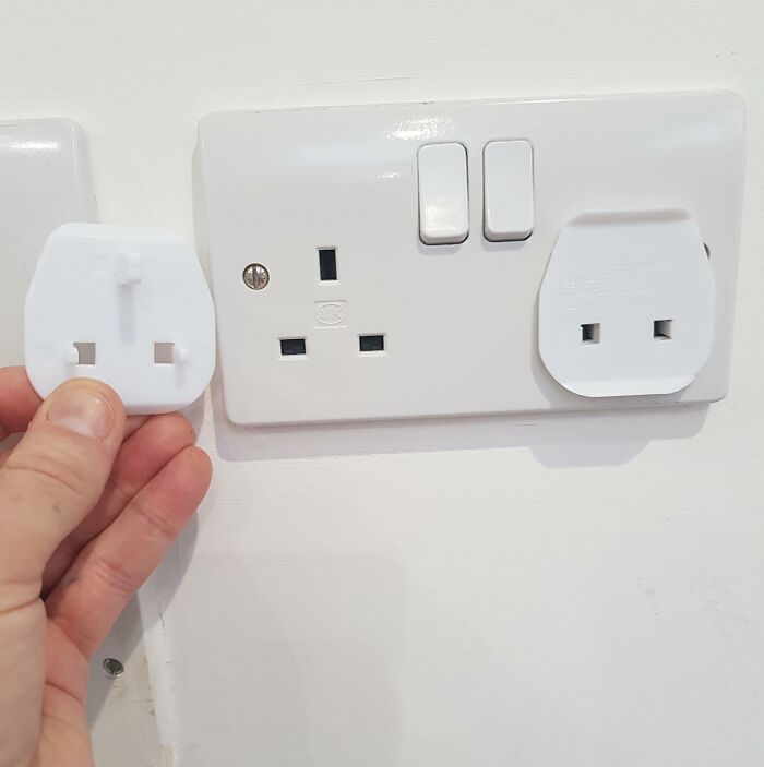 Hand holding a useless plug design next to a UK power socket, illustrating terrible product design that makes no sense.