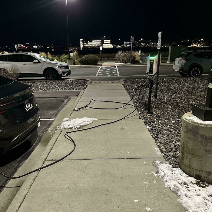 Electric vehicle charging station cable extending across sidewalk, posing a hazard in terrible product design example at night.