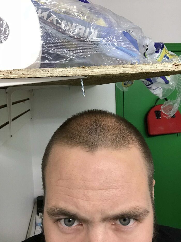 Man with a concerned expression standing under a wooden shelf with a protruding s***w in a reckless work moment.