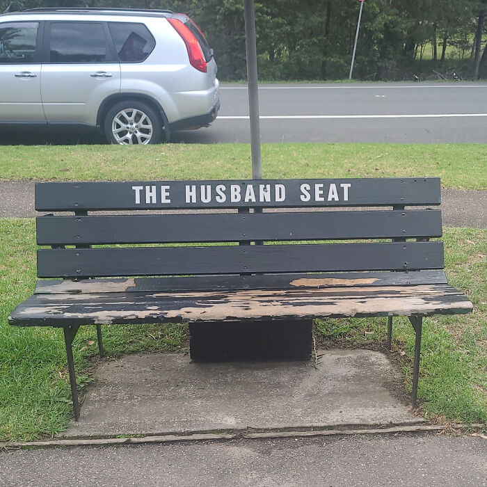 Wooden bench labeled the husband seat, showing gendered nonsense in a public park setting next to a parked silver car.