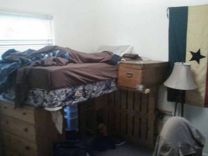 Improvised DIY bed setup with drawers underneath and a makeshift nightstand attached to the bed frame in a small bedroom.