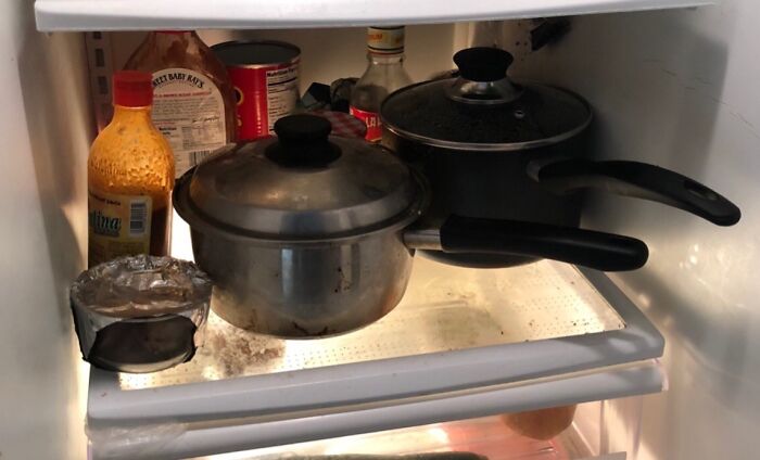 Cluttered fridge shelf with dirty pots, sauces, and containers showing roomates' messy living habits.