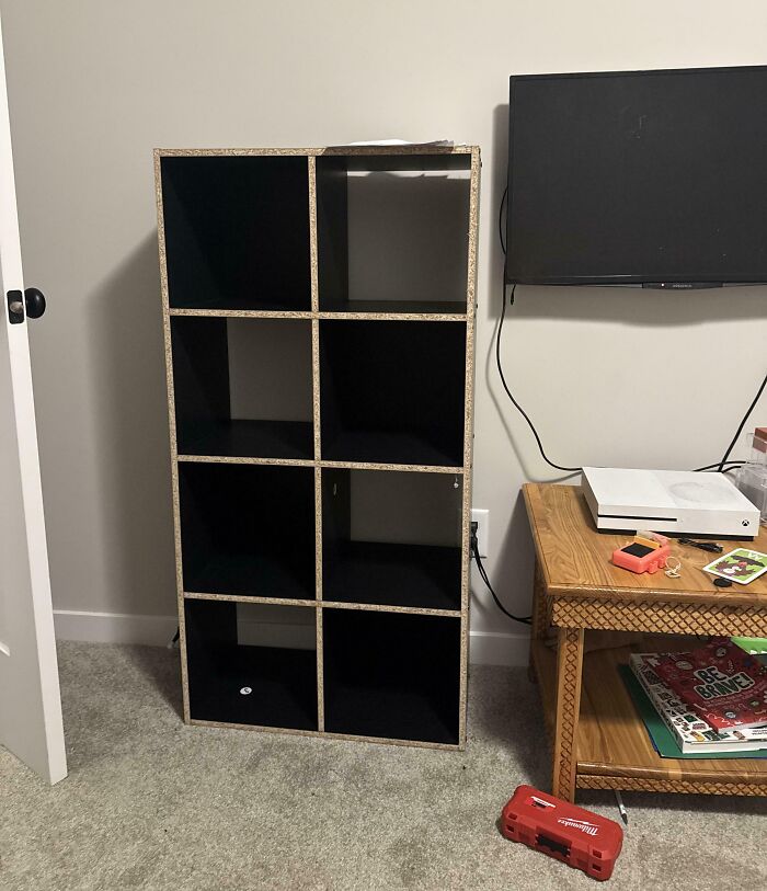 Empty black shelving unit missing its back panel leaning against wall next to TV and wooden table, showing men being men example.