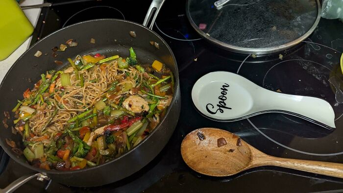Cooking pot with mixed veggies and noodles on stove, wooden spoon and ceramic spoon rest nearby, men being men humor context