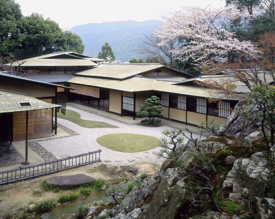 Traditional Japanese architecture featuring low wooden buildings, manicured gardens, and natural mountainous surroundings in spring.