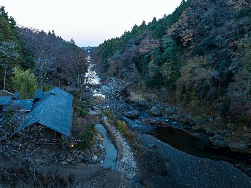 Traditional Japanese architecture nestled beside a forested river valley during early evening in a serene natural setting.