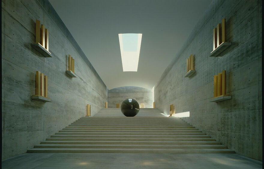 Minimalist Japanese architecture interior with concrete walls, stairs, and a reflective black sphere centered under natural skylight.
