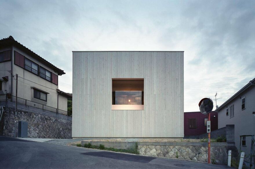 Modern minimalist Japanese architecture with a cubic wooden house featuring a centered window on a sloped street.
