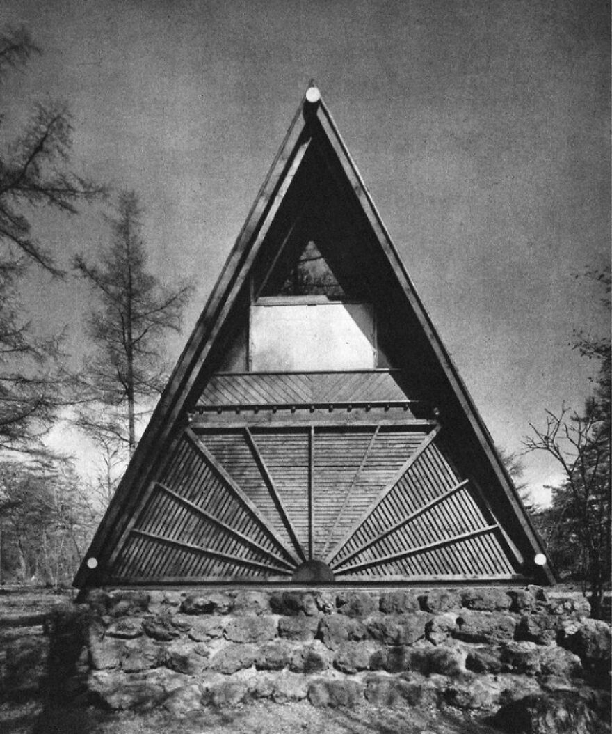 A-frame Japanese architecture example with a triangular roof and wooden slats on a stone foundation surrounded by trees.