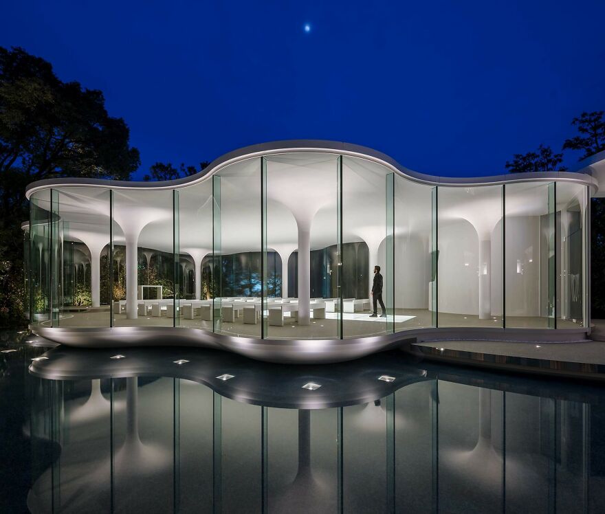 Modern Japanese architecture featuring a glass-walled building with curved roof columns illuminated at night by the water reflection.
