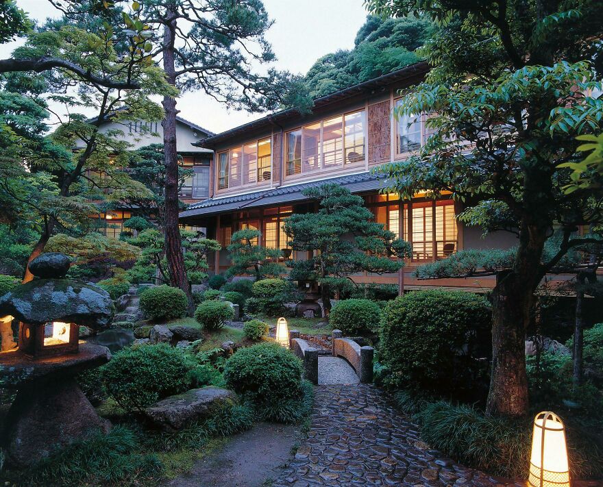 Traditional Japanese architecture with wooden house and serene garden featuring stone lanterns and lush greenery at dusk.