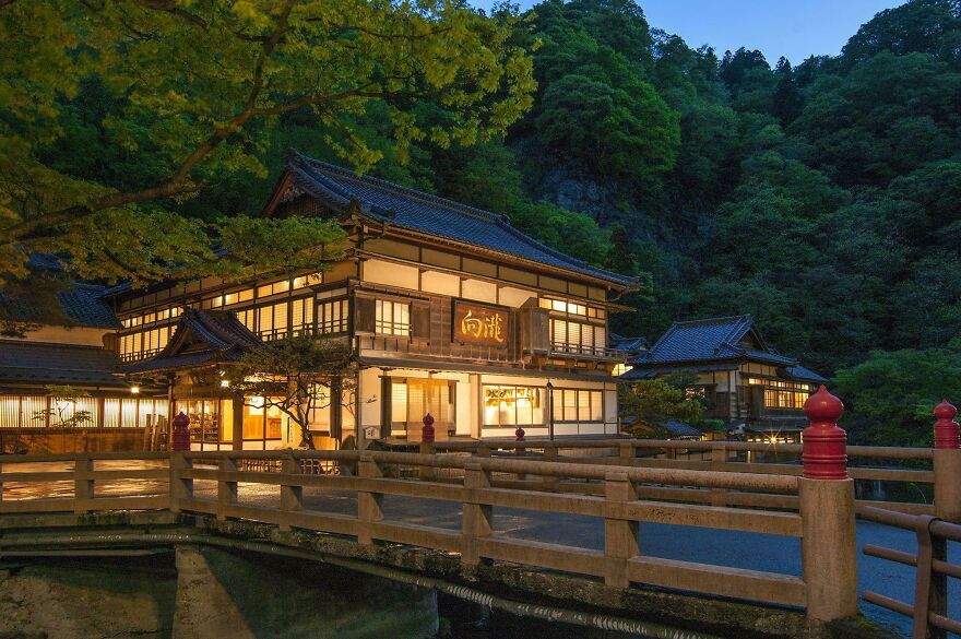Traditional Japanese architecture building illuminated at dusk with wooden bridge and lush green forest in the background.