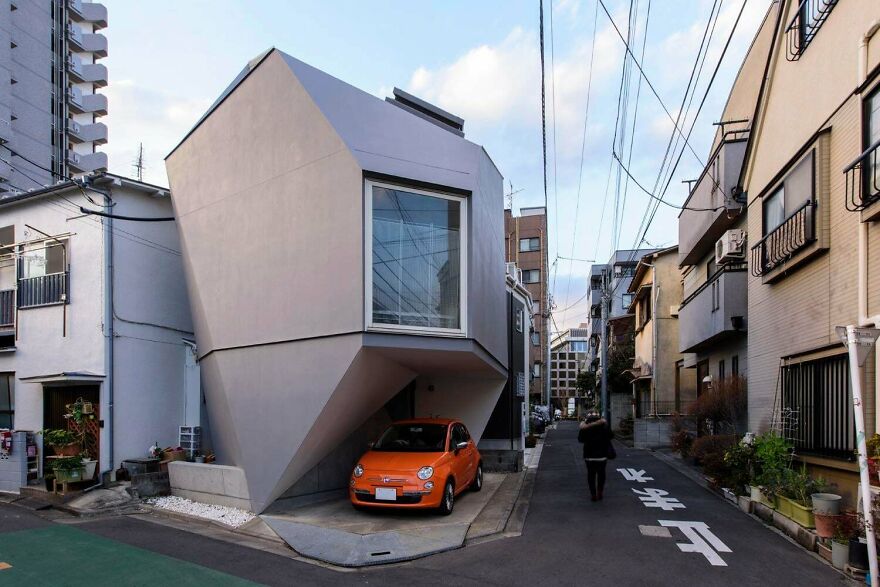 Modern angular Japanese architecture house with a compact orange car parked underneath on a narrow urban street.