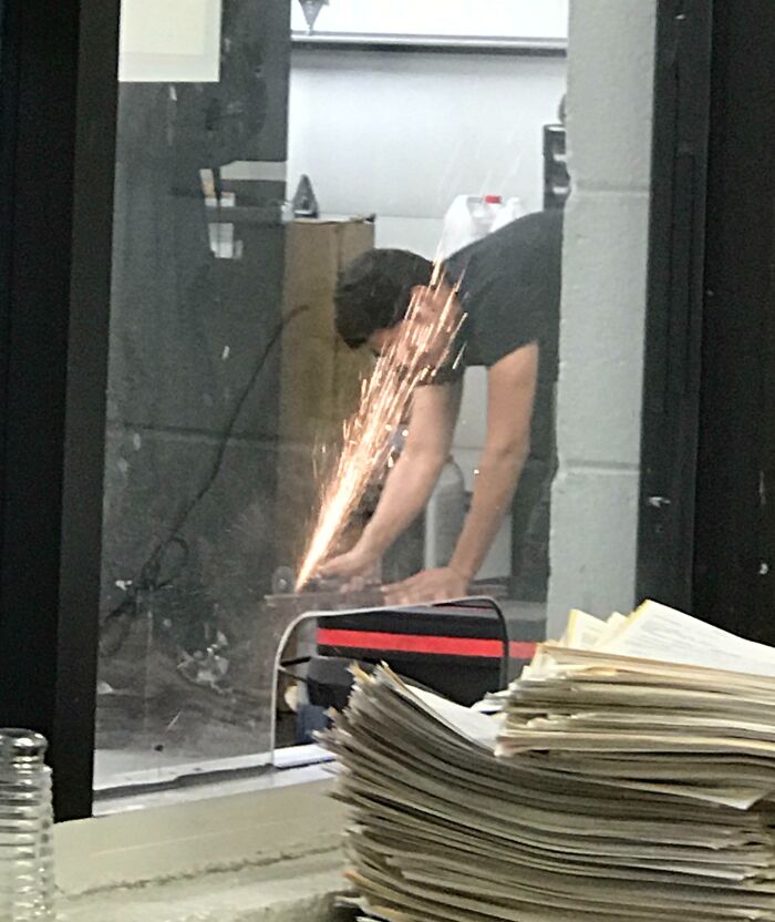 Person grinding metal indoors without safety gear, sparks flying dangerously close to a stack of papers in a reckless work moment.