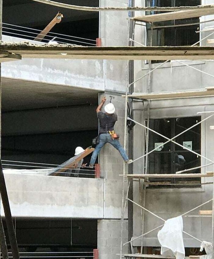 Construction worker hanging recklessly between scaffolding and building without proper safety gear on site.