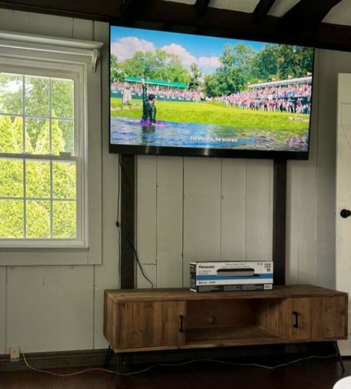 TV mounted too high above wooden media console with box resting on top in a bright room, showcasing infuriating examples of men being men.