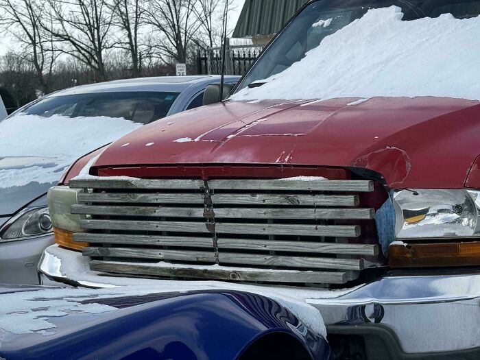 Red truck with a makeshift grill repair made of metal bars covered partially in snow in a parking lot.